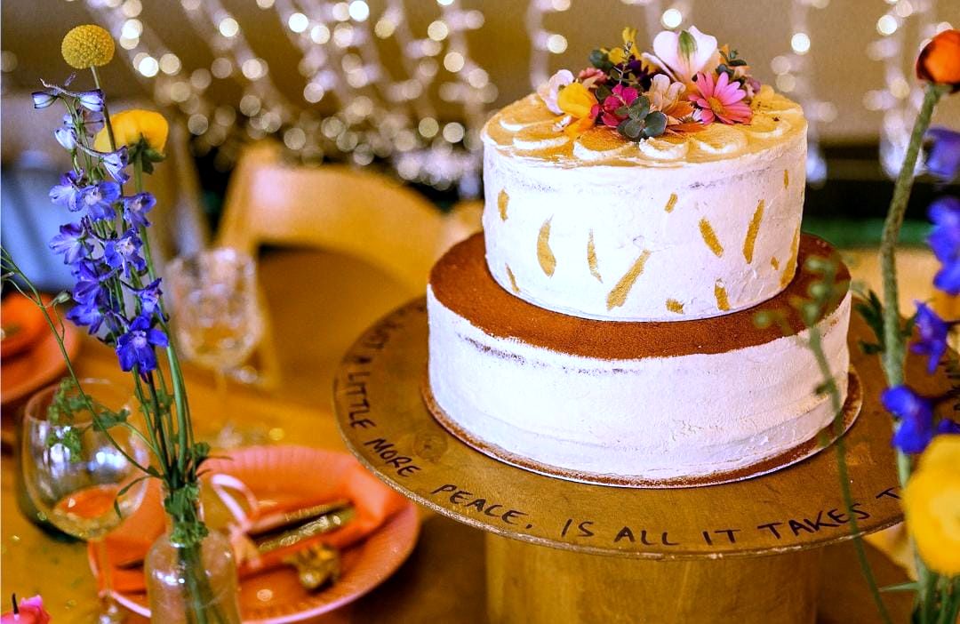 Two-tier semi-naked cake topped with edible flowers on a wooden stand, at an evening party with fairy lights
