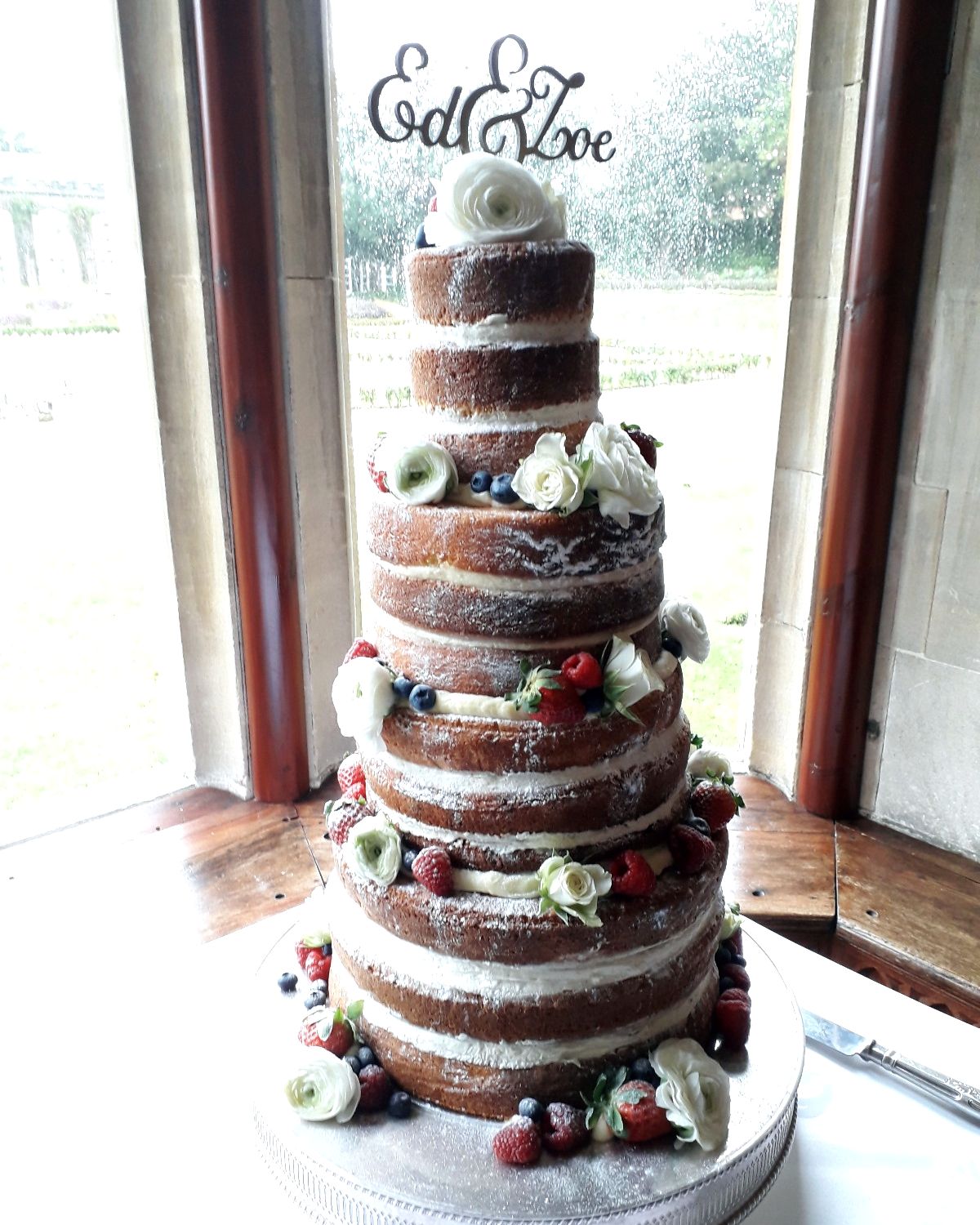 Tall naked wedding cake with white roses and fresh berries in a window