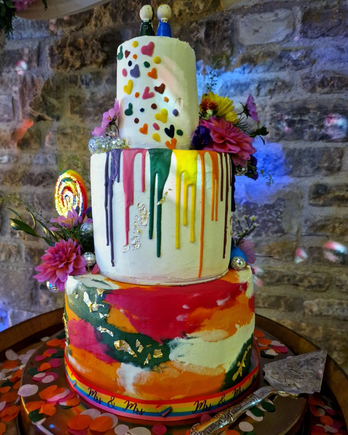 Three-tier wedding cake with rainbow drips and colourful painted base on a barrel